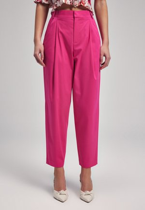 Bright pink high-waisted trousers with pleats, tapered legs, and side pockets. Worn with light-colored, bow-accented heels.