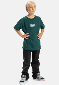 Camiseta de algodón verde con cuello redondo y logotipo blanco, combinada con pantalones negros y zapatillas grises con detalles en naranja y cordones blancos.