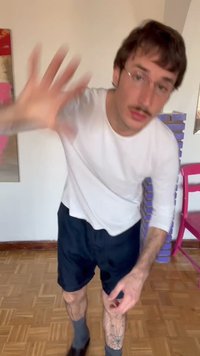 Jonge man met bril die met een wazige hand naar voren reikt, gekleed in een wit overhemd met lange mouwen, donkere korte broek en tatoeages op zijn benen, in een kamer.