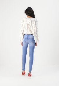 Vit volangblus med långa ärmar i kombination med åtsittande ljusblå jeans. Modellen har röda högklackade skor. Framsidan av designen är inte synlig.