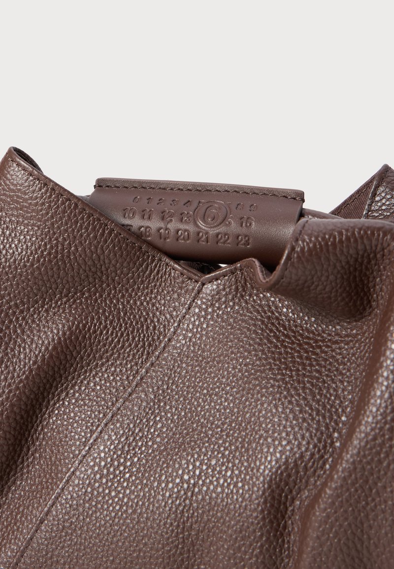 Nahaufnahme einer strukturierten braunen Ledertasche mit einer genähten Lasche, auf der eingeprägte Zahlen und die umkreiste Zahl 6 zu sehen sind.