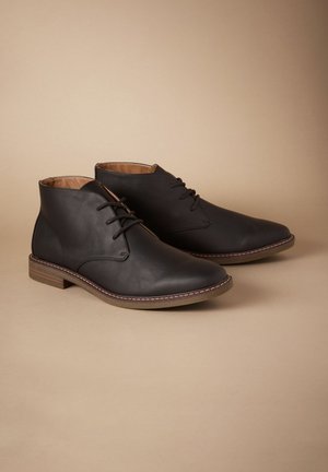 Schwarze Leder-Stiefeletten mit spitzen Zehen, Schnürdesign und hellbraunen Sohlen. Mit dezenten Nahtdetails entlang der Kante.