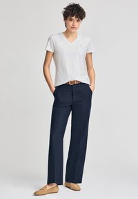 Witte v-hals t-shirt, gecombineerd met marineblauwe broek en een bruine riem. Model draagt tan instap schoenen en staat met de handen in de zakken.