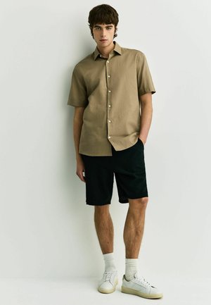 Mladý muž stojící u bílé zdi, oblečený v krátkým hnědém košili na knoflíky, černých šortkách, bílých ponožkách a bílých teniskách.