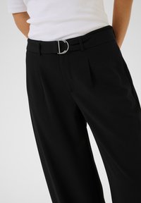 Pantalons larges noirs en tissu lisse, dotés d'une boucle en forme de D argentée sur une ceinture et de plis sur le devant pour un détail supplémentaire.