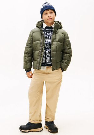 Giacca trapuntata verde oliva, maglione navy a fantasia, camicia crema, pantaloni beige e scarponi da trekking neri indossati da un ragazzo con un berretto lavorato a maglia blu.