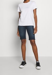 Camiseta blanca con logotipo azul, combinada con pantalones cortos de mezclilla oscura y un ajuste slim. Zapatillas negras con suelas blancas completan el atuendo.