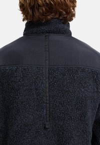 Giacca in pile blu scuro con un corpo testurizzato e morbido e un pannello superiore liscio, caratterizzata da una striscia verticale in nylon lungo il retro.