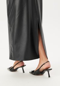 Langer schwarzer Lederrock mit einem seitlichen Schlitz, kombiniert mit schwarzen Slingback-Pumps mit Absatz, die eine Schleifendetail an der Zehe haben.