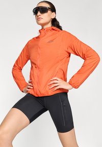 Orange lätt jacka med dragkedja fram, elastiska manschetter och huva, kombinerad med svarta formpassande shorts med sidoficka med dragkedja.