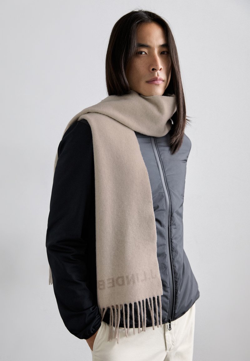 Béžový vlněný šátek s jemnou texturou, s třásněmi na koncích, přehozený přes tmavý kabát. Na šátku se nachází nenápadné logo v světle barevném textu.
