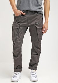 Pantalon cargo en tissu gris foncé. Comprend plusieurs poches, des accents zippés et une coupe décontractée. Porté avec des baskets blanches.