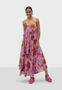 Robe maxi fleurie rose avec des bretelles spaghetti, arborant des motifs vibrants de fleurs et de feuilles, assortie à des sandales à plateforme roses.