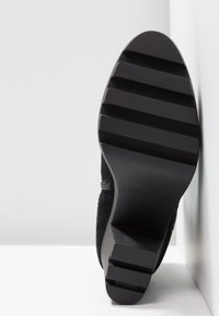 Mustat mokkasiinit, joissa on teksturoitu pinta, esittelevät litteän kumipohjan, jossa on vaakasuorat urat ja kaiverrettu brändäys sivussa.