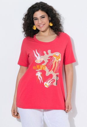 Frau mit lockigem Haar trägt rotes T-Shirt mit gelb-orangefarbenem Quallenmotiv und weißen Hosen, lächelt vor weißem Hintergrund.