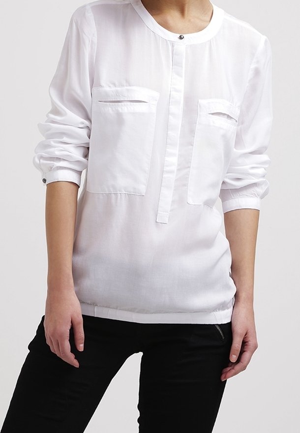 Vit långärmad blus med rund halsringning, två framfickor och knappstängning. Tillverkad av lätt tyg med en mjuk textur.