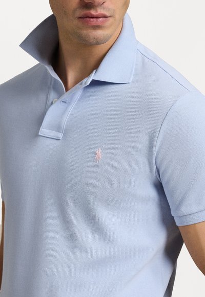 Polo de culoare bleu deschis, dintr-un material texturat, cu plasă cu două butoane și guler înălțat. Prezintă un mic logo brodat roz pe piept.