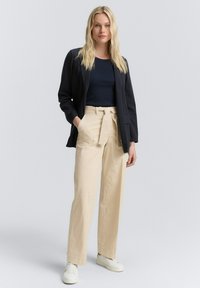 Blazer scuro, top navy e pantaloni beige a vita alta con cintura legata. Sneakers bianche completano l'outfit. Texture morbide nei materiali.