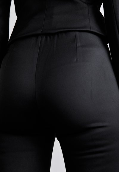Tenue noire sur mesure comprenant un haut ajusté et un pantalon lisse et extensible. Le tissu semble élégant avec un léger éclat et peu de plis.