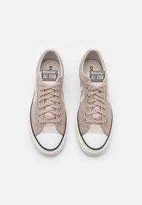 Beige kanva lenkkarit, joissa on valkoinen kuminen varvastoppa ja musta pohja. Varustettu nauhoilla ja "Converse All Star" -merkinnällä kielekkeessä, sekä kyljessä oleva tähti-logo.