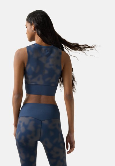 Kvinde med langt, flydende hår iført en ærmeløs marineblå mønstret cropped top og matchende højtaljede leggings, set bagfra på hvid baggrund.