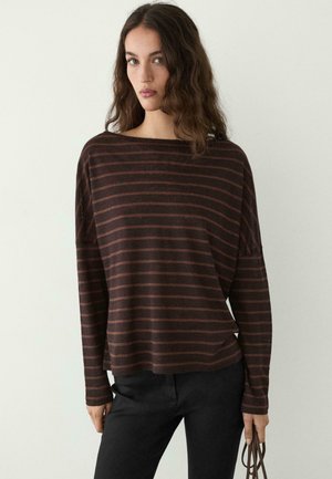 Donna con capelli mossi castani che indossa un top a maniche lunghe largo a righe marroni e nere e pantaloni neri, che tiene una tracolla marrone.