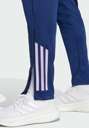 Pantaloni sportivi navy con aperture laterali con cerniera, dotati di tre strisce verticali rosa. Abbinati a scarpe da ginnastica bianche.
