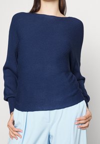 Granatowy sweter z prążkowanego materiału z dekoltem jachtowym i luźnym krojem. Posiada długie rękawy i lekko skrócony dół, zestawiony z jasnoniebieskimi spodniami.