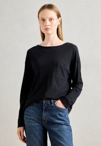 Woman with straight blonde hair wears black long-sleeve top with pocket and blue jeans, standing against a plain light gray background.