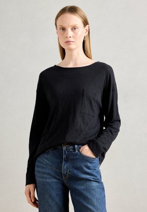 Kvinne med langt, rett blondt hår iført svart langermet topp med lomme og blå jeans, står mot en ensfarget lys grå bakgrunn.