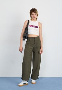White cropped tank top with "RACER" in blue and red lettering, paired with olive green cargo pants featuring pockets, and white sneakers with green accents.