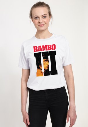 Kvinde med lyst hår iført hvid T-shirt med rød tekst "RAMBO" over stort sort romersk tal II med en mands ansigt indeni.