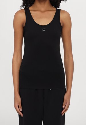 Vrouw draagt een zwarte mouwloze tanktop met een klein vierkant logo in het midden van de borst.