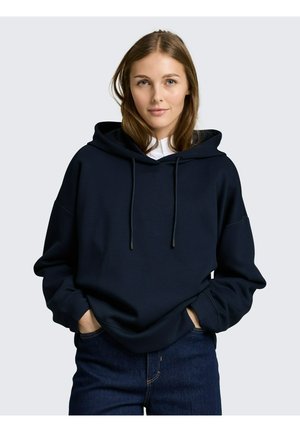 Junge Frau trägt einen marineblauen Kapuzenpullover mit Kordelzügen und dunkelblaue Jeans, Hände in den Taschen, neutraler Hintergrund.