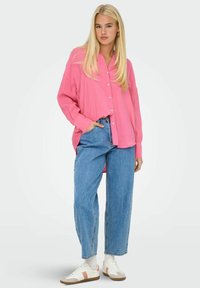 Camicia rosa over-size con colletto, abbinata a jeans a vita alta blu chiaro e sneakers bianche con dettagli rosa.