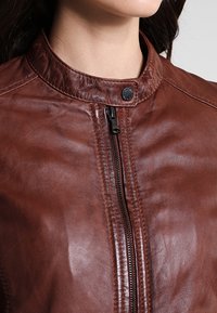 Nahaufnahme einer Person, die eine braune Lederjacke mit einem Reißverschluss vorne und einem Druckknopfkragen trägt, vor einem einfarbigen Hintergrund.