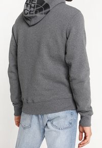 Sudadera gris con capucha que presenta un logo negro en la parte posterior. El material parece suave, con puños y dobladillo de canalé. Combinada con jeans de color azul claro.