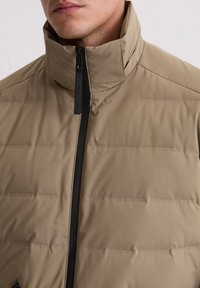 Beige isolierte Jacke mit hohem Kragen, vertikalem Steppmuster und schwarzem Reißverschluss. Verfügt über ein Label am Kragen zur Markenkennzeichnung.