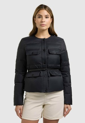 Nő fekete gombos, öves steppelt dzsekit és világos bézs rövidnadrágot visel, és egy egyszerű világosszürke háttér előtt áll.