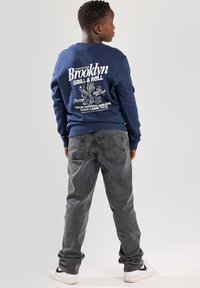 Sudadera azul marino con un gráfico de "Brooklyn Grill & Roll" en la parte posterior, combinada con jeans grises y zapatillas blancas, vista desde atrás.