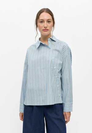 Femme portant une chemise à rayures bleu clair avec des boutons sur les côtés, associée à un pantalon bleu foncé, debout devant un fond blanc uni.