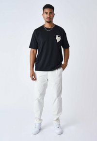 Camiseta negra de manga corta con un diseño de corazón en el lado izquierdo del pecho, emparejada con pantalones blancos y zapatillas blancas. Estilo simple y moderno.