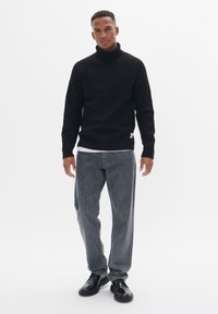 Suéter negro de canalé con cuello alto y mangas largas, combinado con jeans grises y zapatos negros. Camiseta blanca sencilla visible.