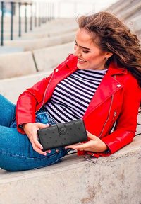 Black wallet with textured dots, rectangular shape, clasp closure, and logo. Model wears a red leather jacket and striped shirt, seated.