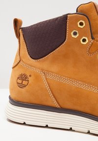 Botte en cuir marron avec semelle en caoutchouc blanche, panneau rembourré noir à la cheville, œillets dorés et logo d'arbre embossé sur le talon.