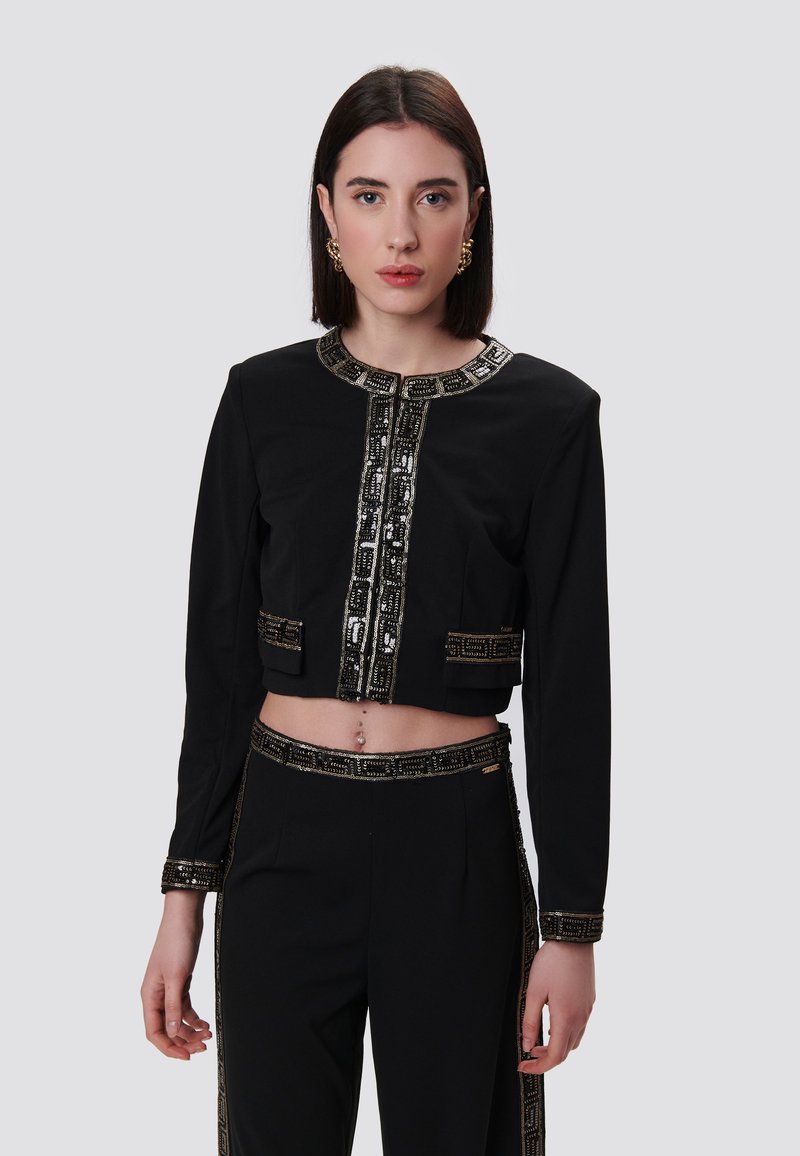 Zwarte cropjack met ingewikkelde gouden en witte versieringen langs de kraag en voorkant, met lange mouwen en voorzakken.