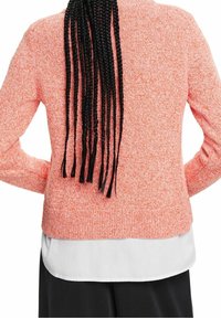 Korallinen neulepusero, jossa teksturoitu pinta, pitkät hihat ja pyöreä pääntie, kerrostettuna valkoisen paidan ja tummien housujen päälle.