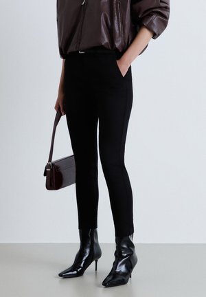Personne portant un pantalon noir ajusté, des bottines noires à bout pointu et talons, un haut marron à manches longues et tenant un sac à main marron.