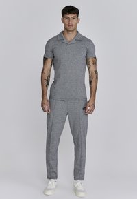 Šedé texturované polokošile s límečkem, doplněná odpovídajícími zúženými kalhotami. Bílé tenisky dodávají outfitu celkový vzhled. Tetování viditelná na pažích.