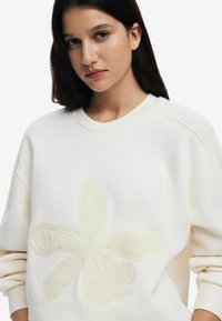 Cream sweatshirt featuring a plush, textured floral design in the center, ribbed shoulder accents, and a rounded neckline.